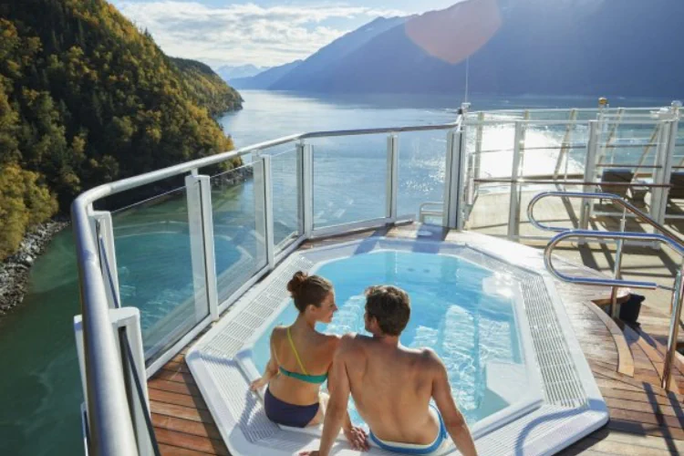Gemini hat gesagtEin Paar entspannt in einem Whirlpool auf dem Deck eines Kreuzfahrtschiffes mit Blick auf eine bergige Küstenlandschaft.