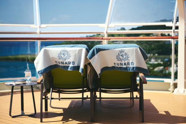 Zwei Sonnenliegen mit Cunard-Handtüchern stehen auf einem Schiffsdeck, daneben ein Beistelltisch mit Drink, im Hintergrund das Meer.